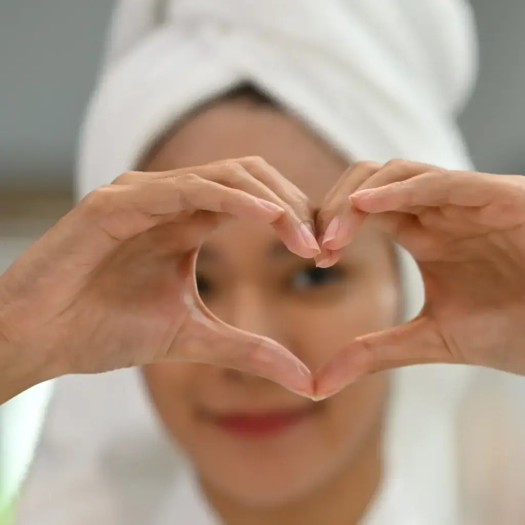 אישה עם מגבת על הראש יוצרת לב עם הידיים כחלק משגרת טיפוח פנים ביתית