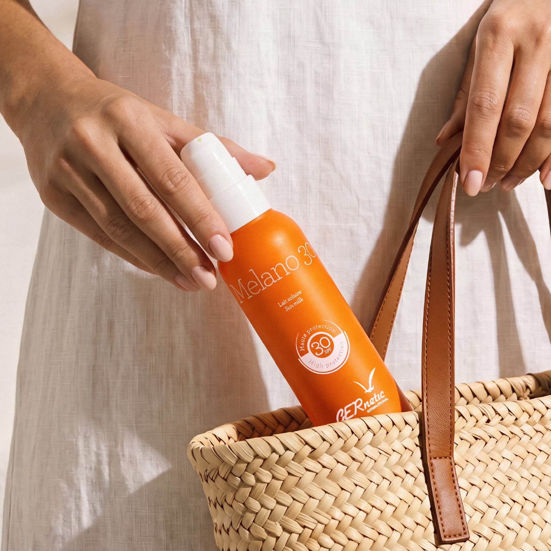 Melano 30 - תחליב הגנה לפנים ולגוף SPF 30; יד אוחזת בבקבוק מלאנו 30 סמוך לתיק קש